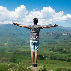 Man standing on a cliff edge with his arms outstretched, overlooking a vast green valley and mountains, symbolizing freedom or success.